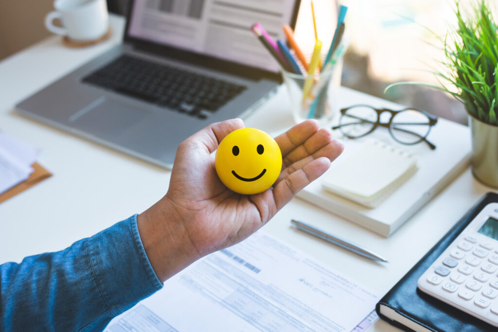 Mental health in the workplace. Person holding a yellow ball with a smiley face in front of their desk.